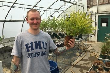 温室里的男学生拿着一株植物. 他的t恤上写着UNH研究生院.